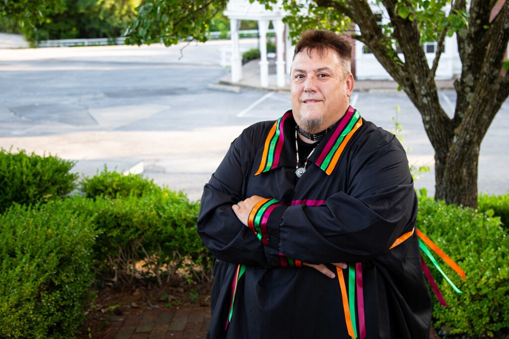 Jay Kelley stands with arms folded across his chest outside. He is wearing a traditional black ribbon shirt with pink, green and orange ribbons on the collar and cuffs.
