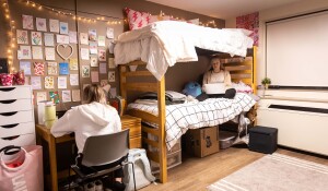 Two female students in sitting in a room in Clinton Hall. One girl is sitting crosslegged on the lower bunk with a laptop on here lap. The other is sitting at the desk.