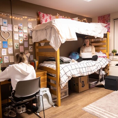 Two female students in sitting in a room in Clinton Hall. One girl is sitting crosslegged on the lower bunk with a laptop on here lap. The other is sitting at the desk.