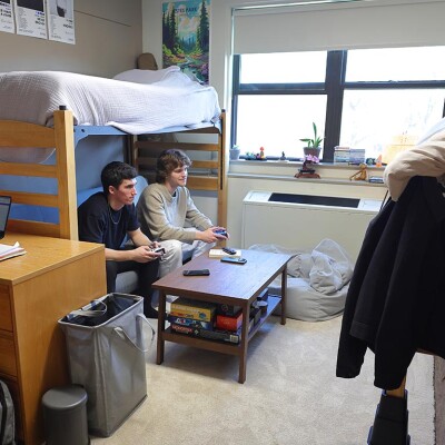 Two male students sit under their bed, which has been raised on risers. They are playing video games.