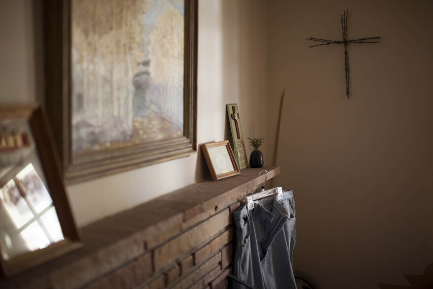 A large picture hangs above a mantle with other photos on the mantle. A pair of jeans hangs from the mantle and a cross hangs on the wall.