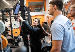 Patricia Dietz points to an EliteForm screen attached to a weight rack while two students look on.