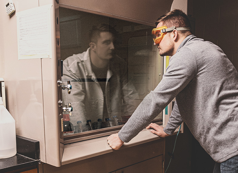 Owen Grover, wearing street clothes, looks into a reflective hood in a science laboratory. The reflection is him in a lab coat.