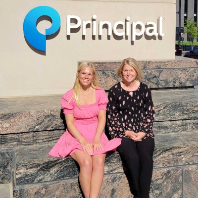 Natalie Thiesfeld ’27 and Jen Bye Christy ’95 sit on a stone wall in front of the Principal sign